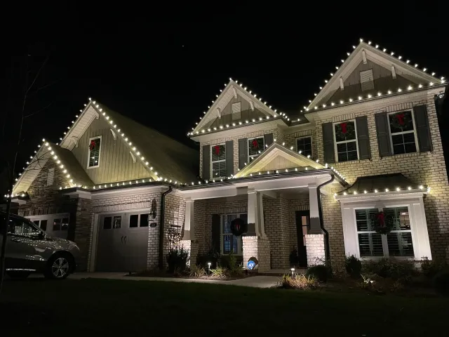 Two-story brick house decorated with white Christmas lights on roof edges and green wreaths with red bows on windows and the front door at night.