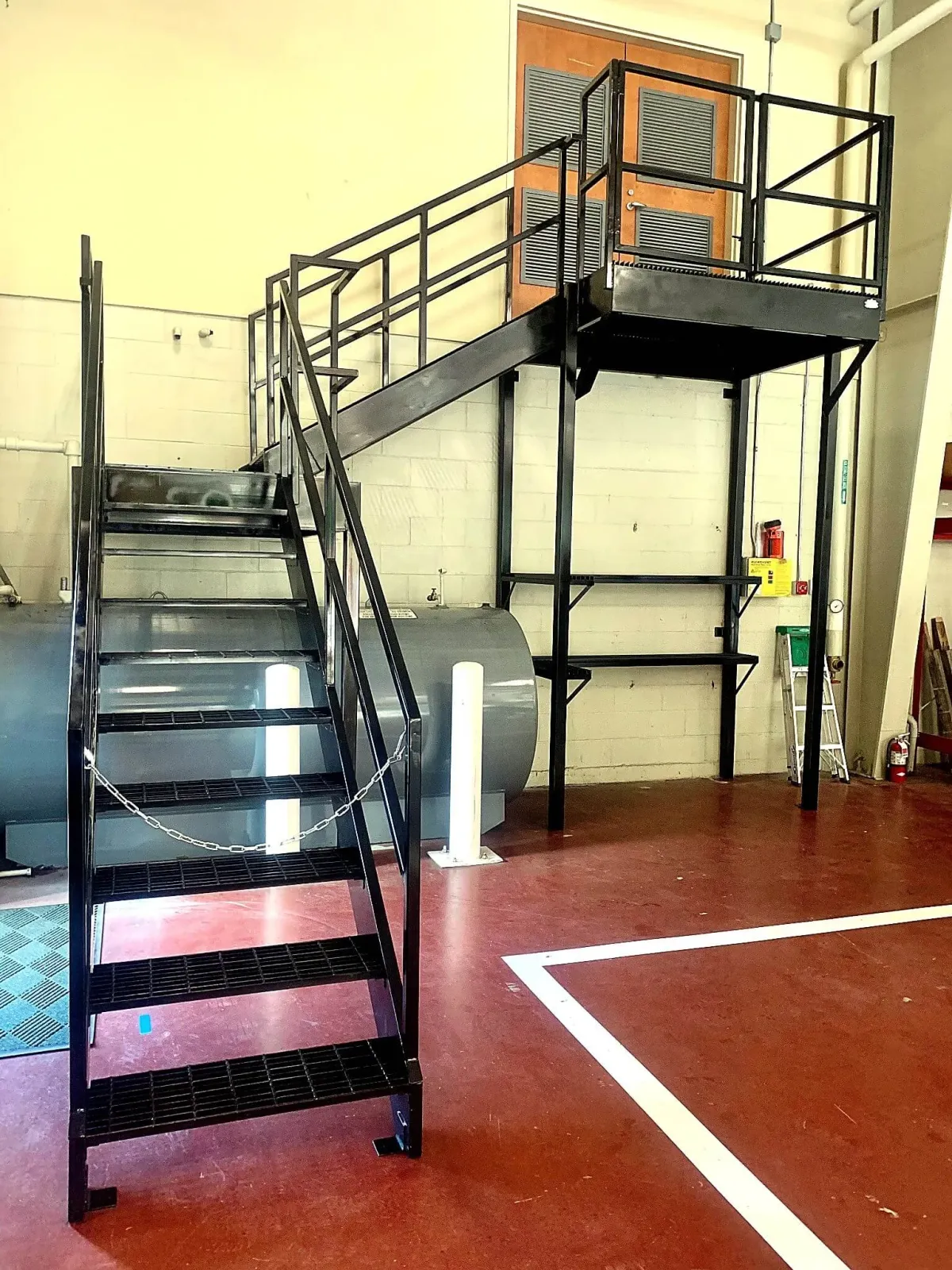 A metal staircase with black railings in an indoor setting. There is a large propane tank underneath the stairs, and a white chain blocking access to the bottom of the staircase. The background includes a wall with a yellowish color, a wooden door, a