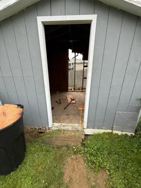 Gray shed with open doorway, showing a dark interior and a dirt path leading to it from the grass.