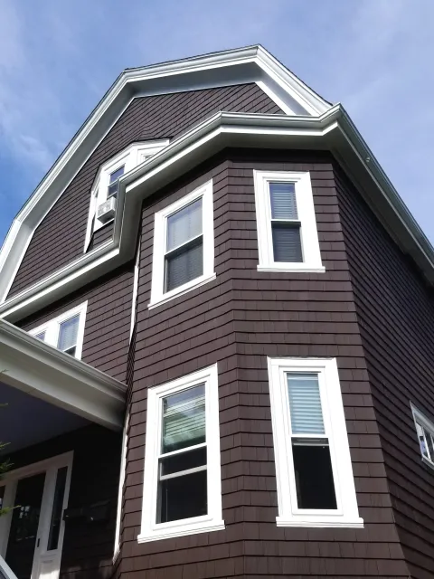 Close-up view of a dark brown house on Jones Hill, Dorchester with white window frames and shingle siding, looking up at the corner of the building against a blue sky.