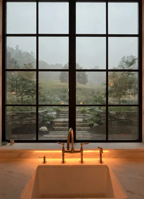 View from a kitchen window across stone garden walls and central steps toward a planted hillside in the rain