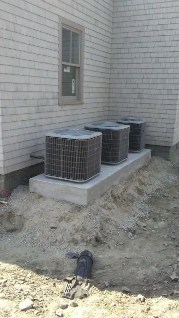Three air conditioning units on a concrete pad outside a building with weathered siding and a window.