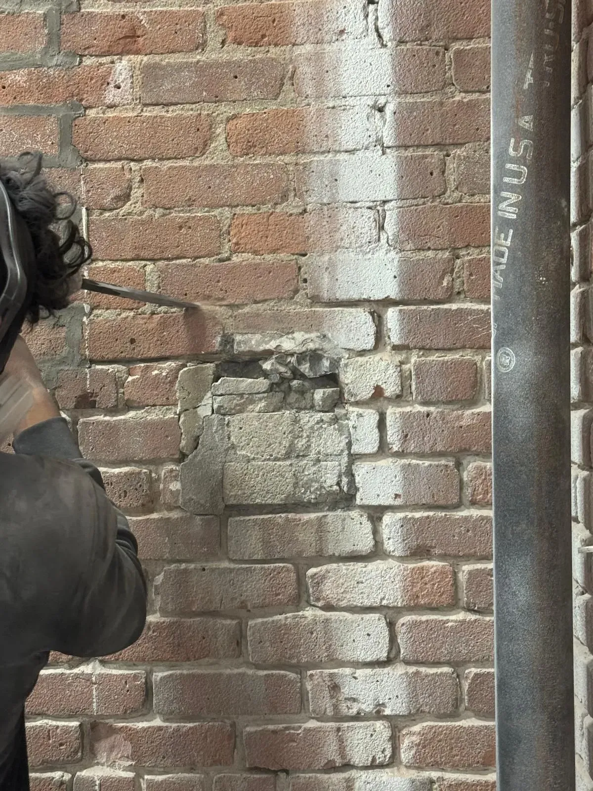 A person using a tool to chip away at a section of a brick wall beside a metal pipe.