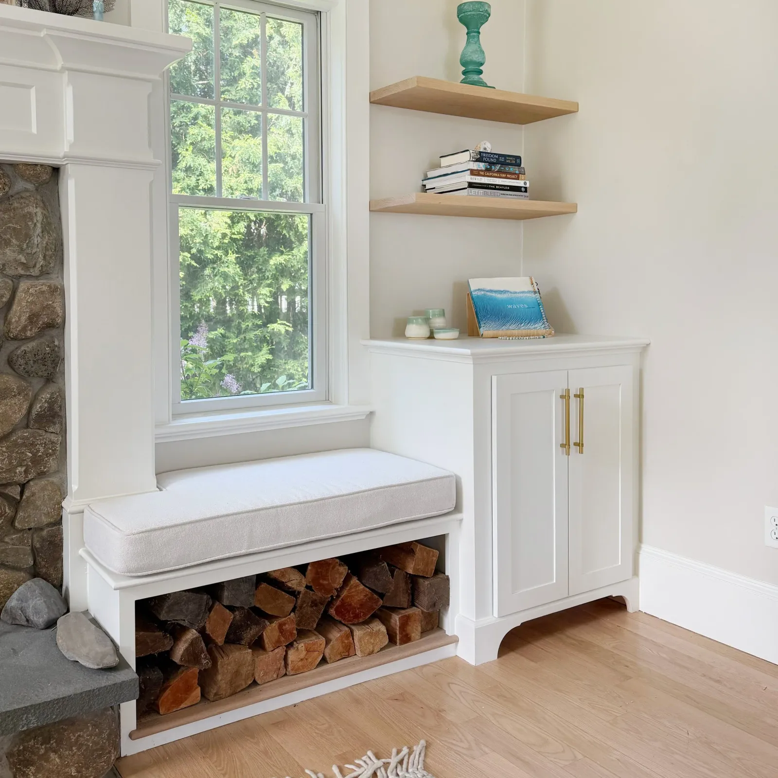 A built-in white bench with storage underneath, a window with a view of trees, two floating wooden shelves with books and decorative items, and a wood-burning stove with stones and firewood underneath.