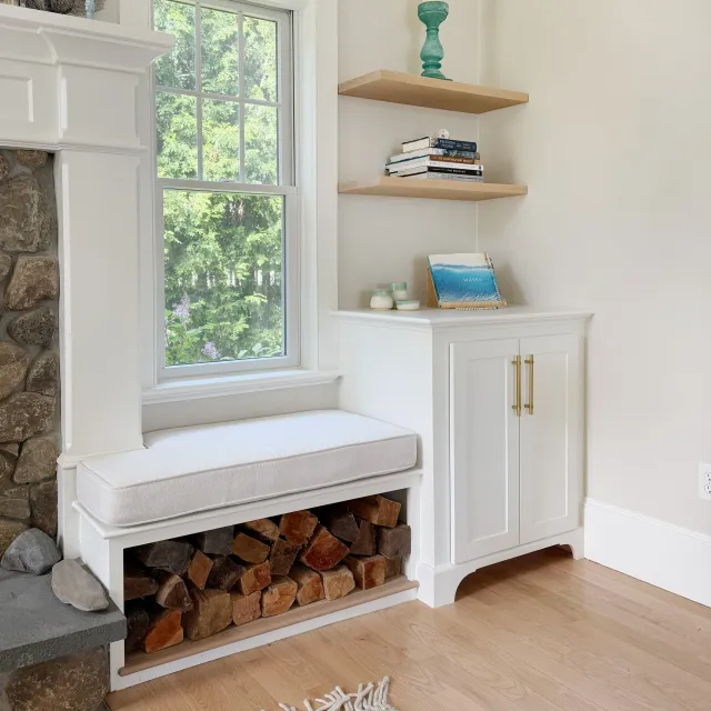 A built-in white bench with storage underneath, a window with a view of trees, two floating wooden shelves with books and decorative items, and a wood-burning stove with stones and firewood underneath.