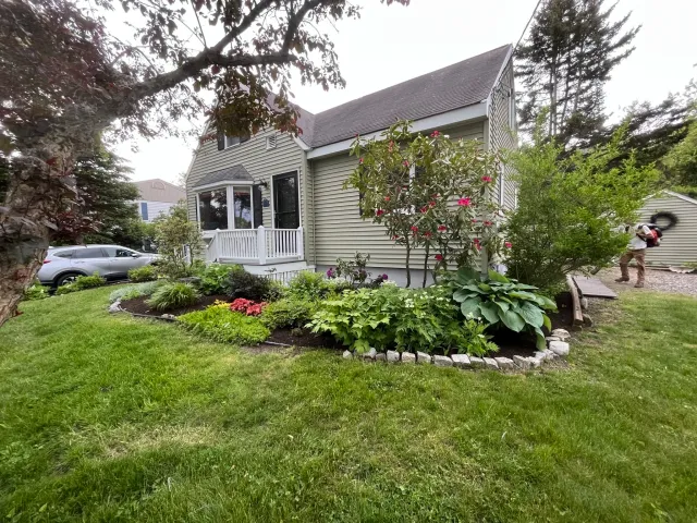 A house with a well-maintained front yard featuring various plants and flowers, a small porch with white railing, a driveway with a parked silver car, and a person walking in the background.