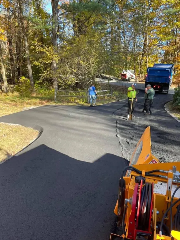 Workers paving a driveway in a wooded setting, with a dump truck and construction equipment visible.