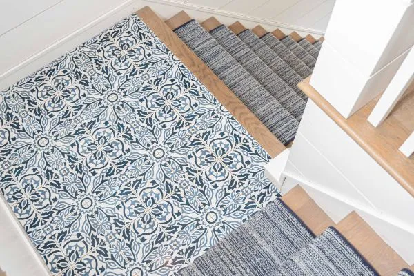 A stylish stair runner installation featuring patterned blue and white tiles at the landing, transitioning to a gray striped runner on the wooden stairs. Part of a stair runner installation project.