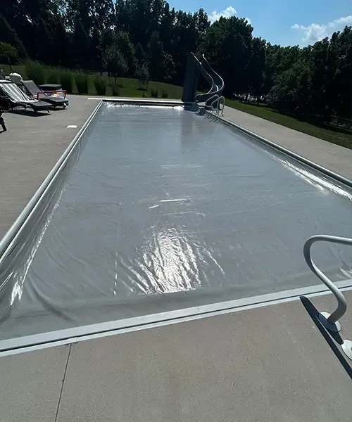 A rectangular outdoor swimming pool is covered with a silver safety cover. Lounge chairs and a table sit on the pool deck, and trees and grass surround the area under a sunny sky.