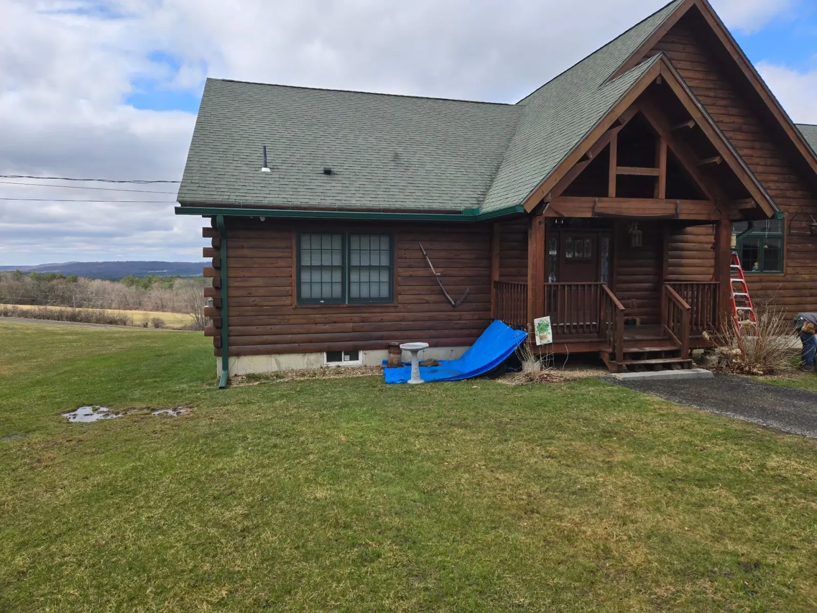 Log cabin with a green roof on a grassy yard, with a blue slide by the porch under a cloudy sky