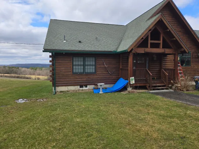 Log cabin with a green roof on a grassy yard, with a blue slide by the porch under a cloudy sky