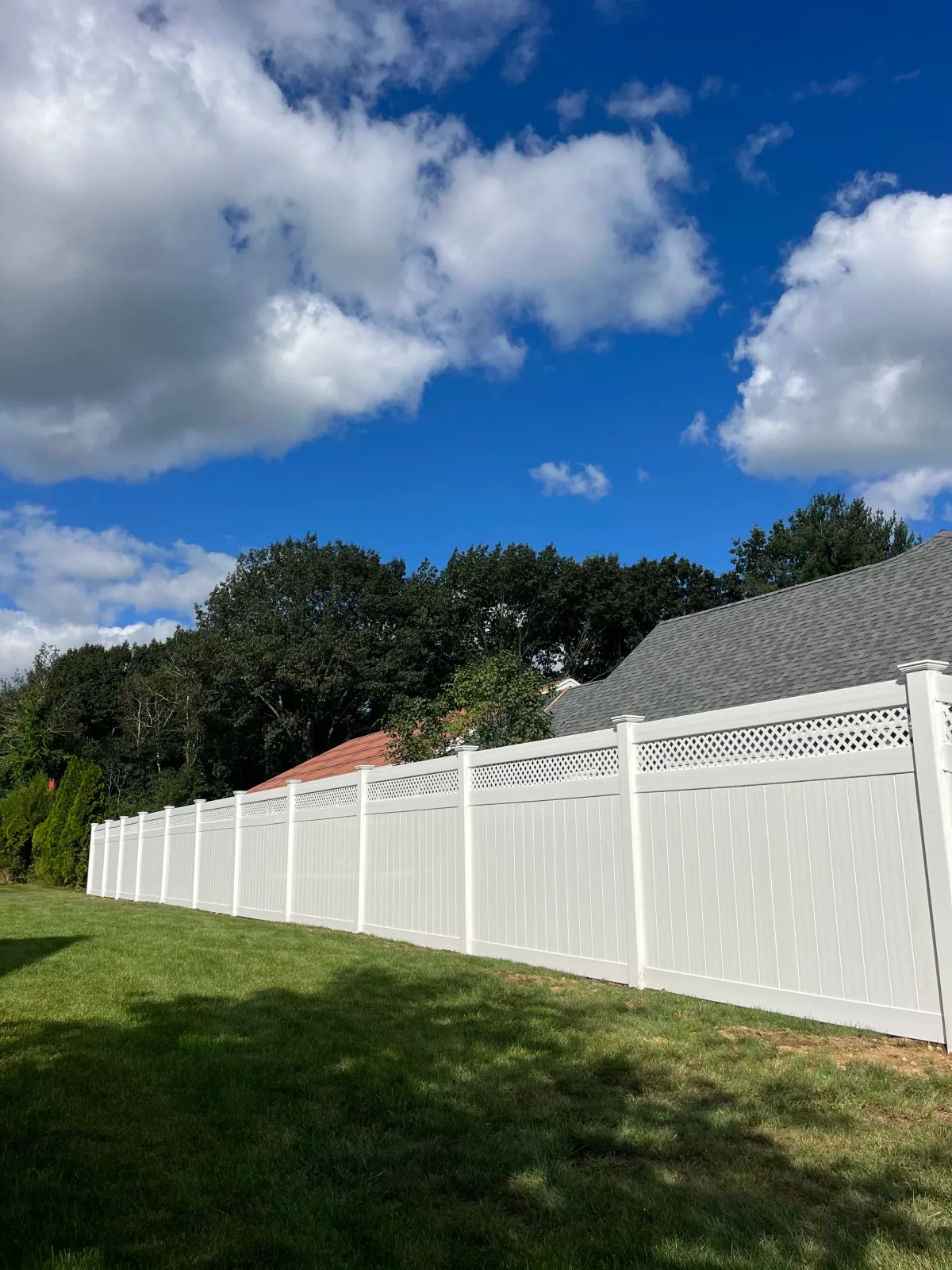 A backyard scene with a white privacy fence, green grass, trees, and a house with a gray shingled roof. The sky is partly cloudy with blue sky visible.