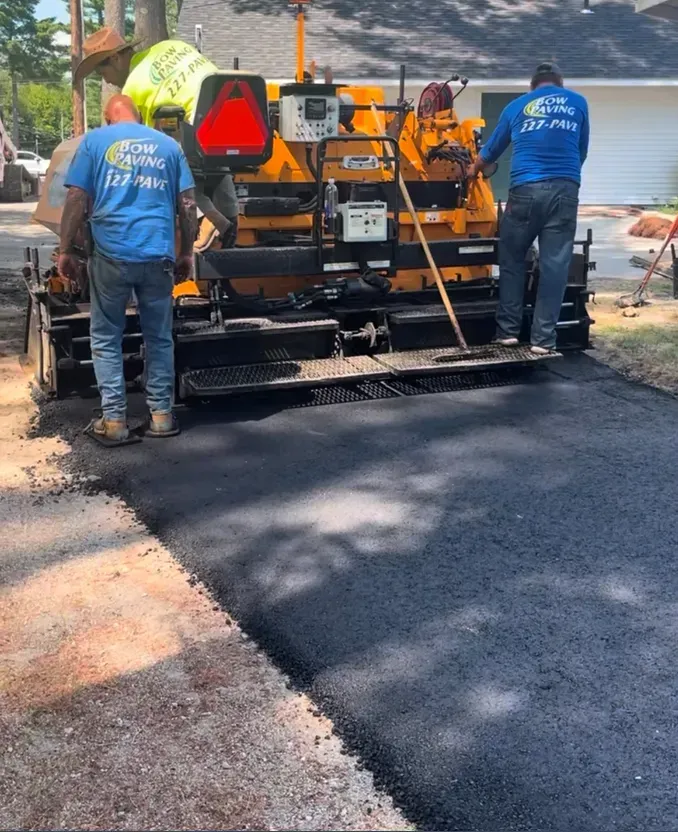 Three workers in blue shirts operate an orange asphalt paving machine, laying a new blacktop driveway in a residential yard.