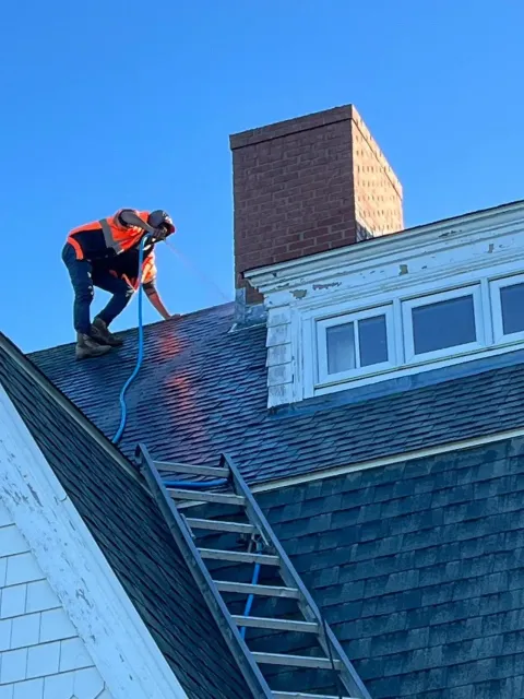 Worker on a steep roof securing a ladder beside a brick chimney and white dormer windows.