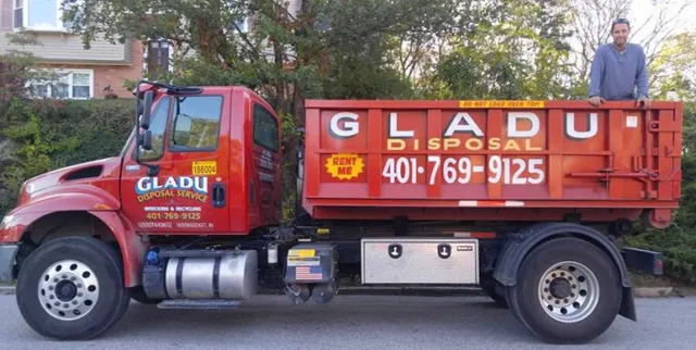 A red disposal truck for Glad U Disposal, with a man standing on top, parked on a street in front of greenery and residential buildings.