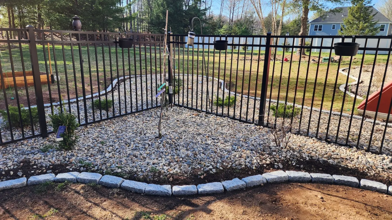 Black metal fence encloses a landscaped garden bed filled with gravel, small plants, and a young tree.