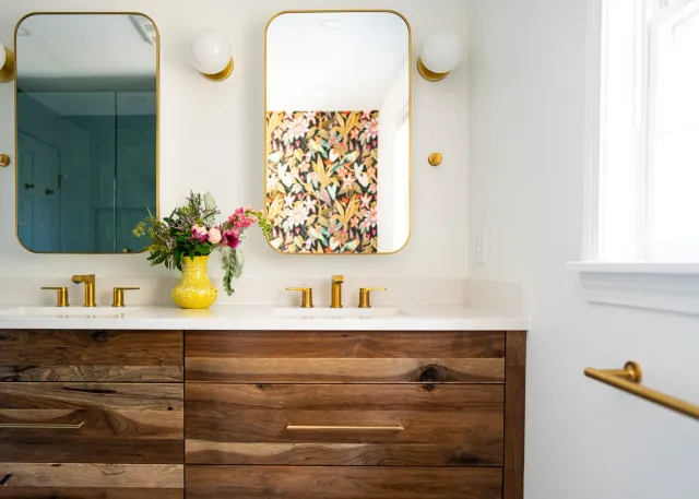 Bathroom vanity with a wooden cabinet, white countertop, two mirrored cabinets with gold trim, two spherical wall sconces, a yellow vase with pink and purple flowers, and gold faucets. There is a window with white blinds and a towel bar on the right.