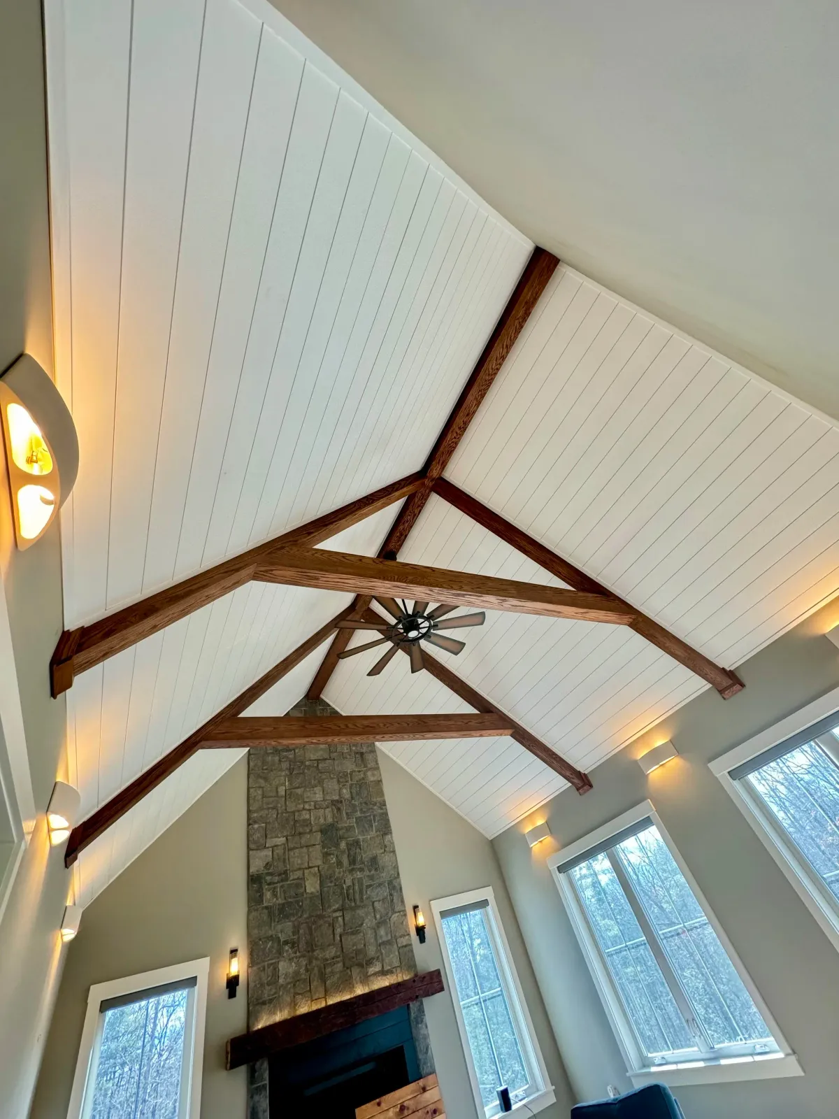 Interior view of a room with a vaulted ceiling with exposed wooden beams, a ceiling fan, a stone fireplace, and large windows.