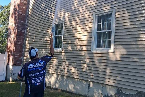 Person in uniform pressure-washing beige vinyl siding of a house, spraying upward near two windows and a brick chimney.