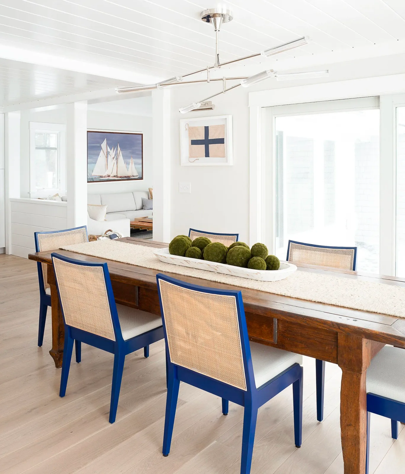 Hardwood floor installation in a Scituate dining area with a wooden table, blue chairs, a white table runner with moss ball decor, large glass sliding doors, and nautical artwork on the wall.