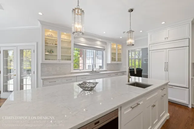 Modern white kitchen with marble countertops, glass-front cabinets, and pendant lighting.