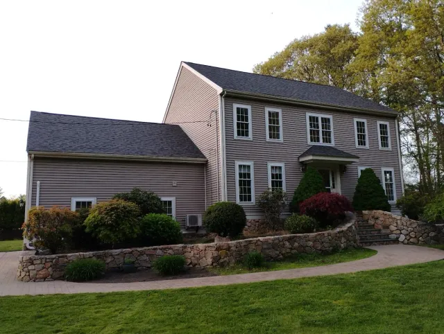 Two-story house with gray siding, dark roof, and landscaping including bushes, a stone wall, and green grass.