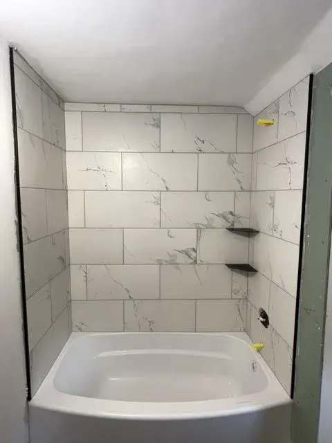A bathroom shower with white marble tiles, two black shelves, and a white bathtub.