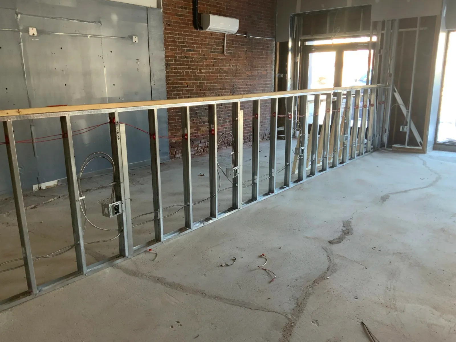 Interior under construction with metal stud wall framing, exposed brick wall, and incomplete electrical work.