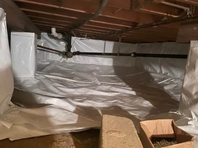 Crawlspace lined with white vapor barrier beneath exposed wooden joists, with a small opening in the floor.