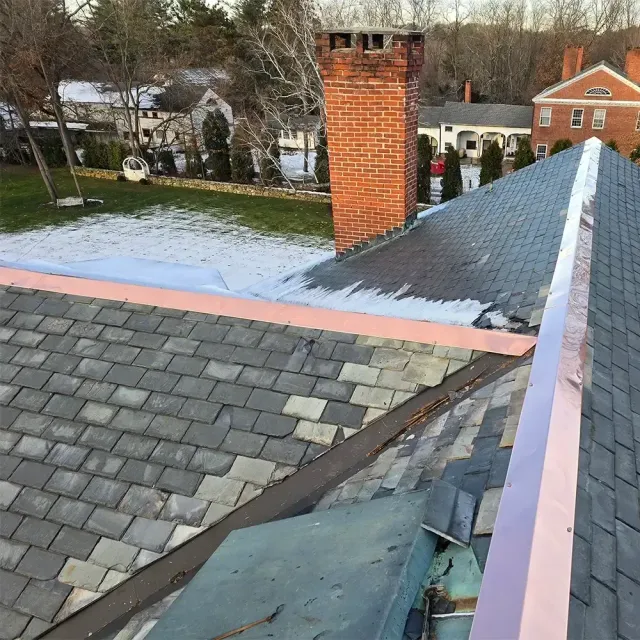 A high-angle view of a slate roof undergoing repairs, featuring new copper flashing and an old brick chimney.