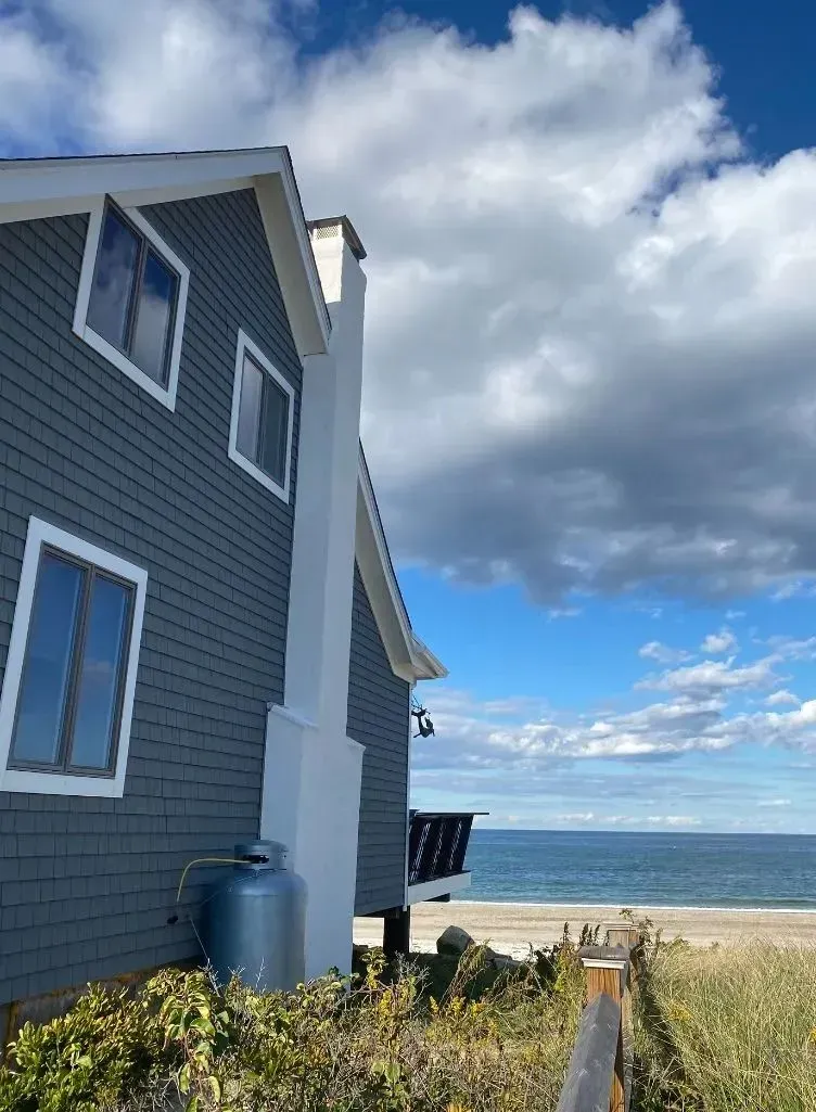 A low-angle view of a grey-shingled beach house with a white chimney, overlooking the ocean under a cloudy blue sky.