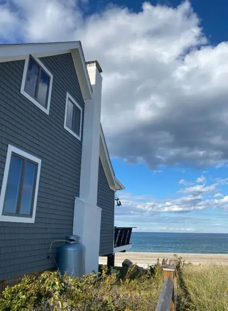 A low-angle view of a grey-shingled beach house with a white chimney, overlooking the ocean under a cloudy blue sky.