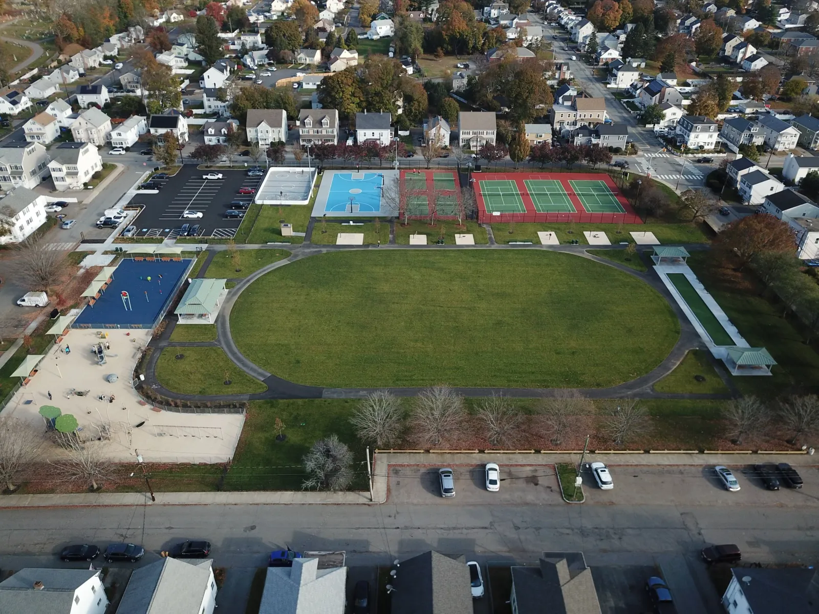 Aerial view of a park with a large oval green field, surrounding running track, multiple sports courts including basketball, tennis, and pickleball, a playground area, a parking lot, and nearby residential houses.