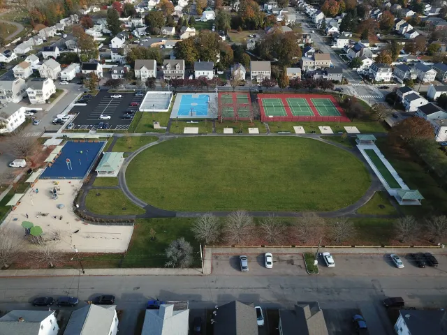 Aerial view of a park with a large oval green field, surrounding running track, multiple sports courts including basketball, tennis, and pickleball, a playground area, a parking lot, and nearby residential houses.