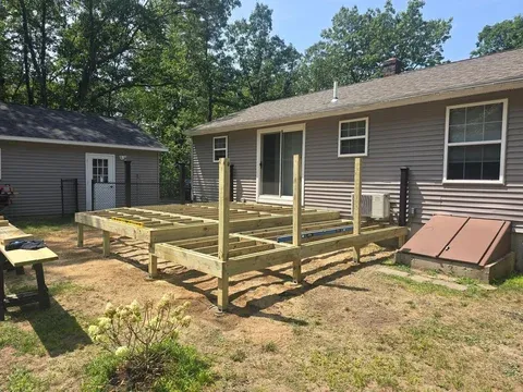 Backyard deck construction in progress with wooden frame and posts, attached to a beige house with small storage shed nearby