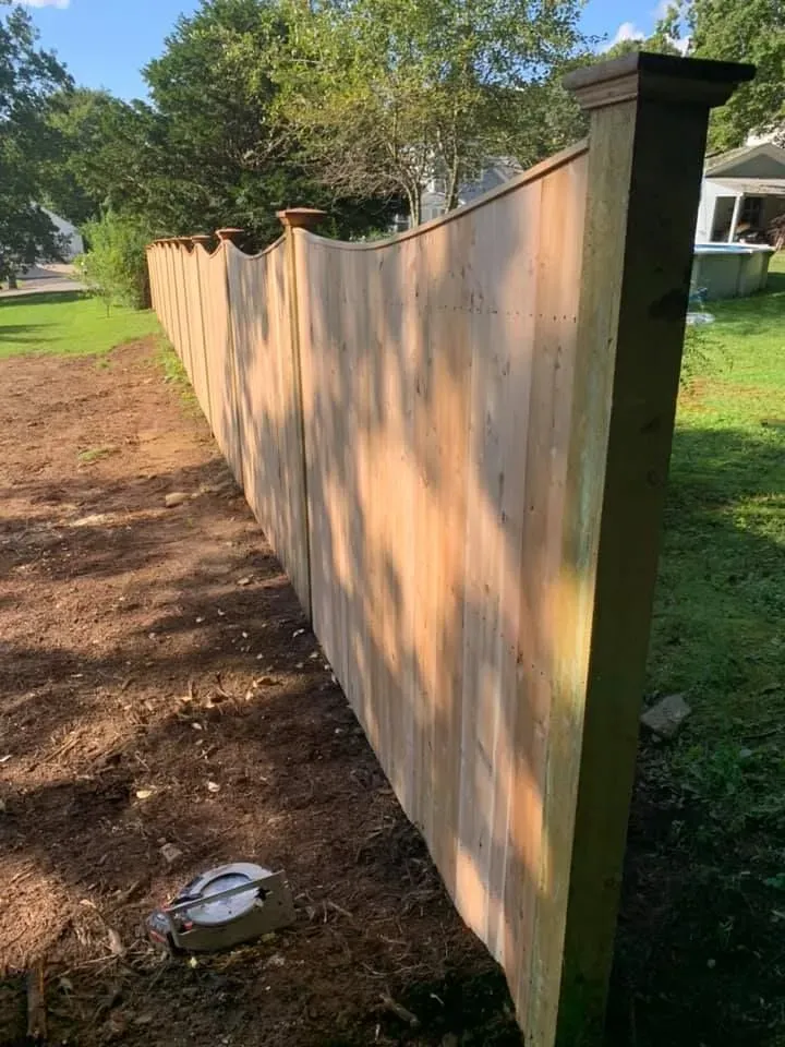 A side-angle view of a newly installed wooden privacy fence with a scalloped top edge in a grassy, suburban yard.