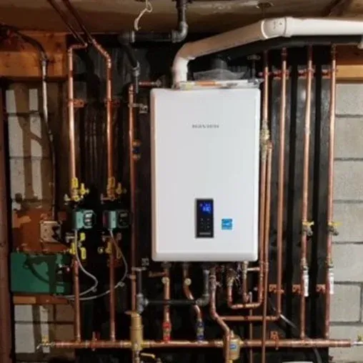 A wall-mounted tankless water heater with multiple copper pipes connected to it, installed in a basement with a concrete wall and exposed wood framing.