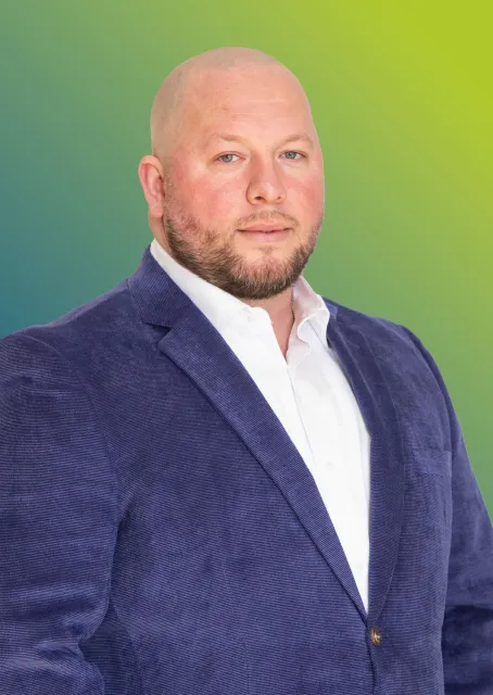 A man with a shaved head and beard, wearing a white shirt and textured blue blazer against a green gradient background.