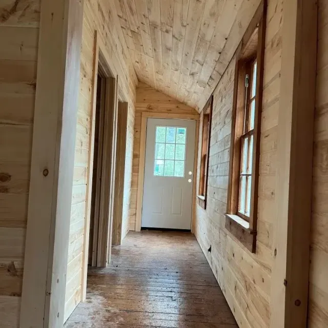 Narrow unfinished wooden hallway with plank walls, floor, and a white door at the end.