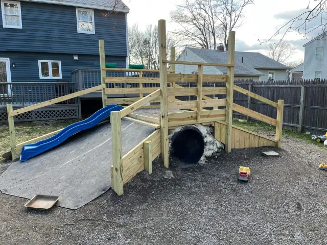 A wooden playground structure with a slide, ramp, and tunnel in a backyard, surrounded by a wooden fence and neighboring houses.