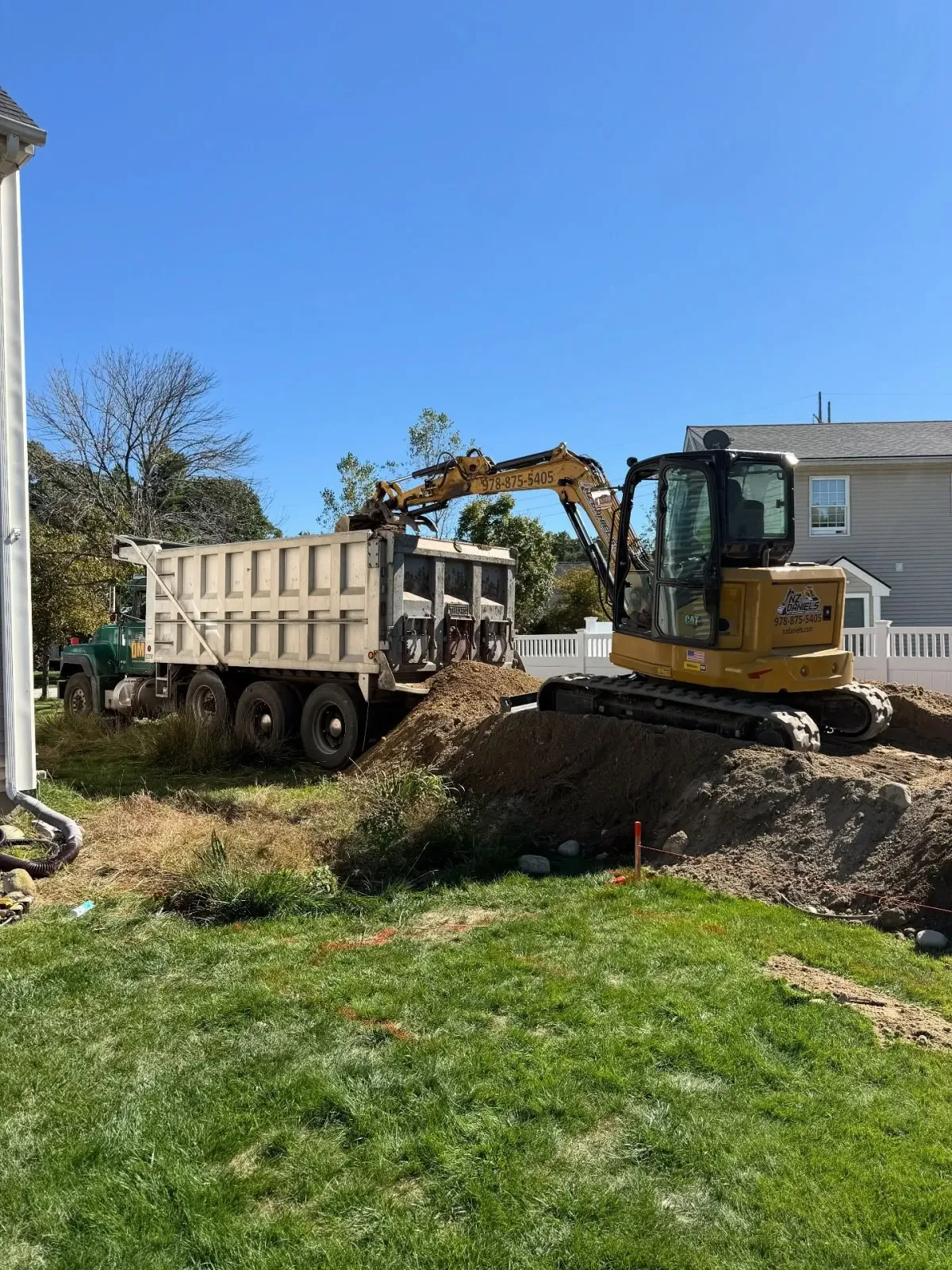 Yellow excavator loading a dump truck with dirt on a construction site next to a house under a blue sky.