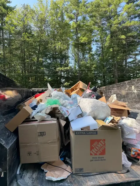 Overflowing dumpster with cardboard and trash bags in a wooded outdoor area