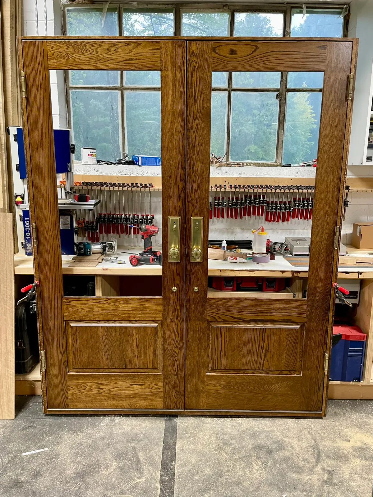 Wooden cabinet with glass panel doors in a woodworking shop, with clamps and tools on the workbench behind it.
