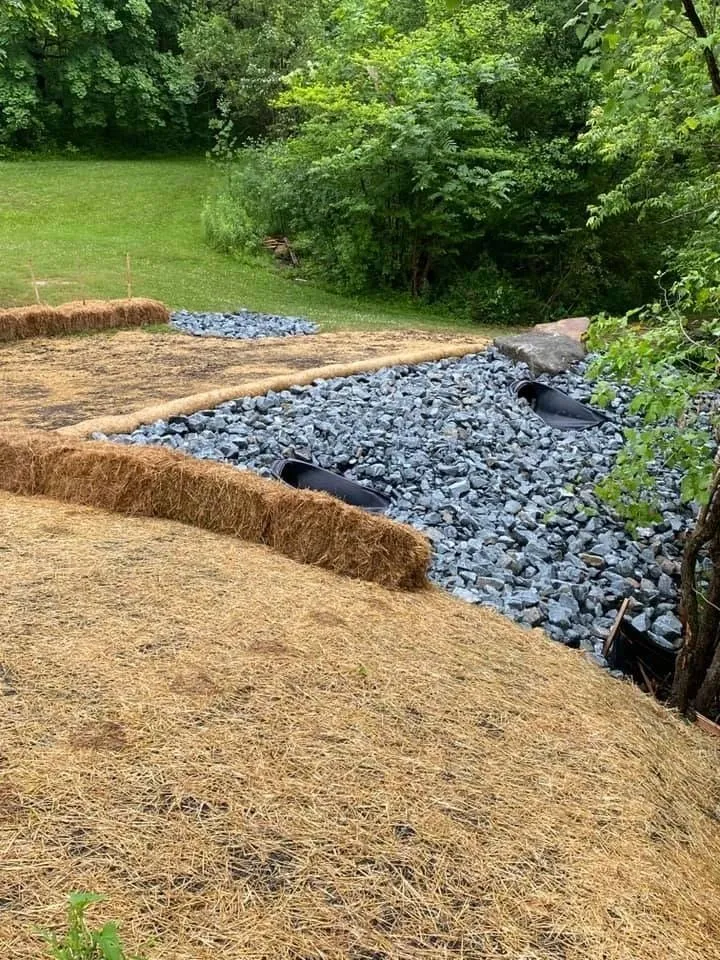 Hay bales and rocks border a channel, with two black pipes and surrounding greenery.