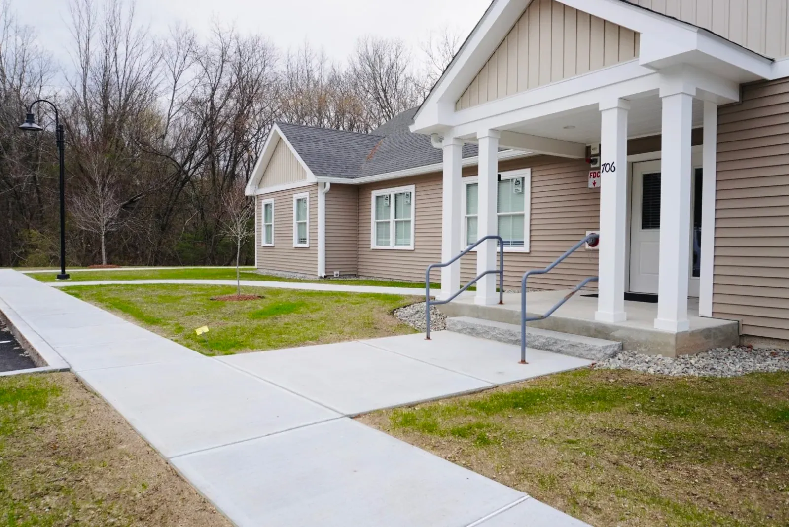 A modern single-story building with tan siding and white trim, featuring a covered entrance with pillars. Concrete walkways and stairs with railings lead to the entrance. The surrounding area includes a small grass lawn with young trees and a streetlamp, bordered by a wooded area.