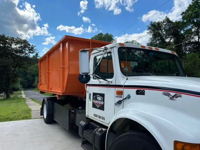 Orange dumpster on a white truck on a residential street under a blue sky with clouds.