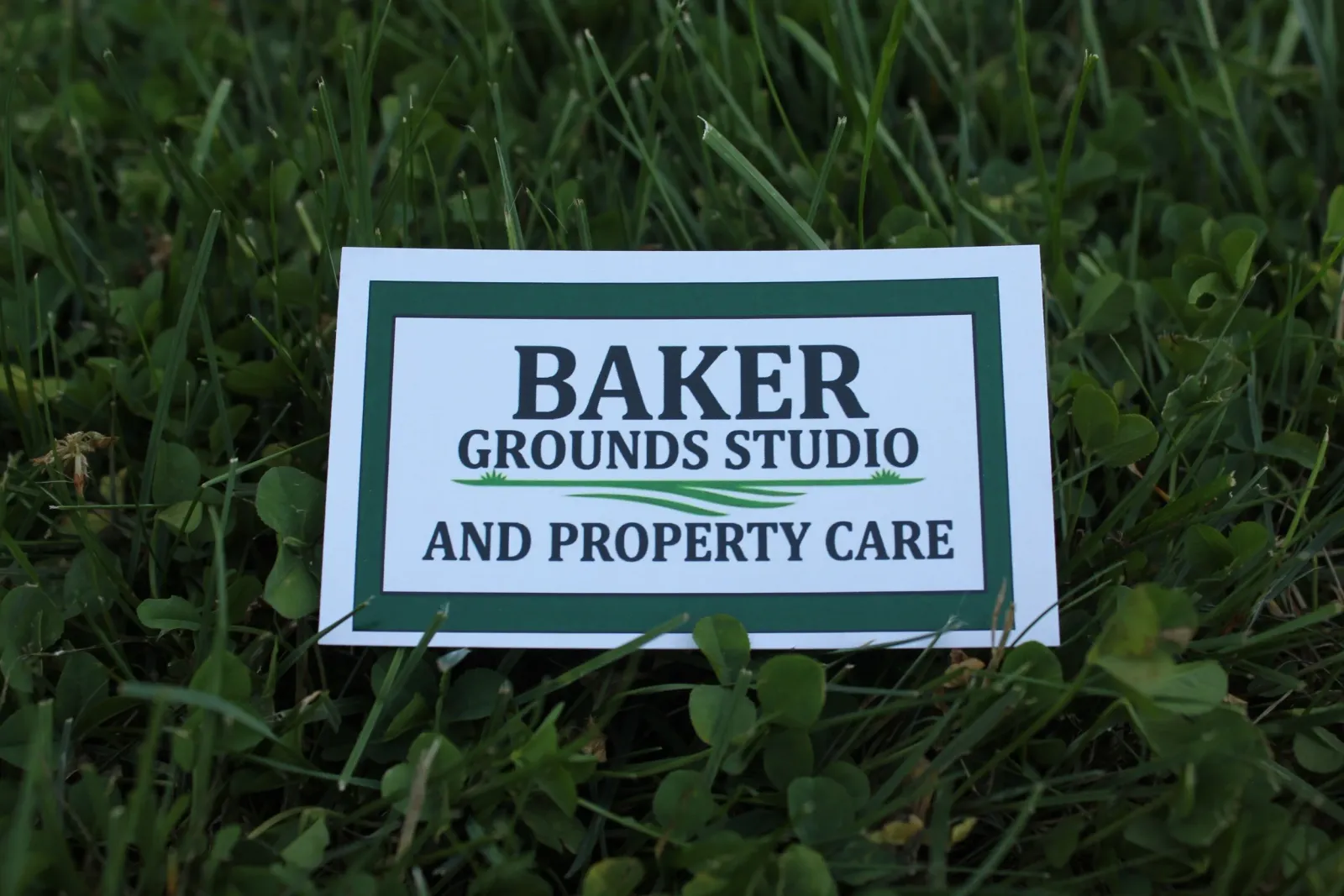 A sign on the ground reads 'Baker Grounds Studio and Property Care', placed amidst green grass and clover plants.