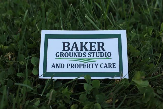 A sign on the ground reads 'Baker Grounds Studio and Property Care', placed amidst green grass and clover plants.