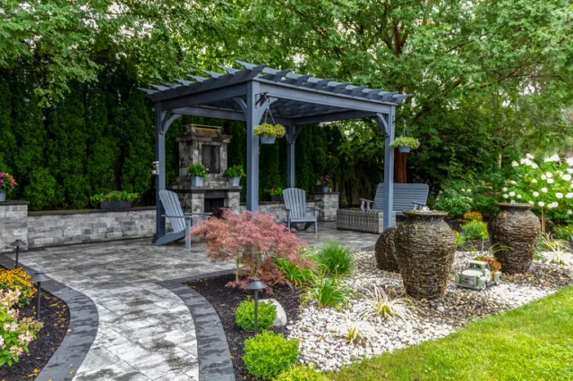 Large gray pergola on pavers with stone fireplace and waterfall urns for backyard oasis landscape architecture