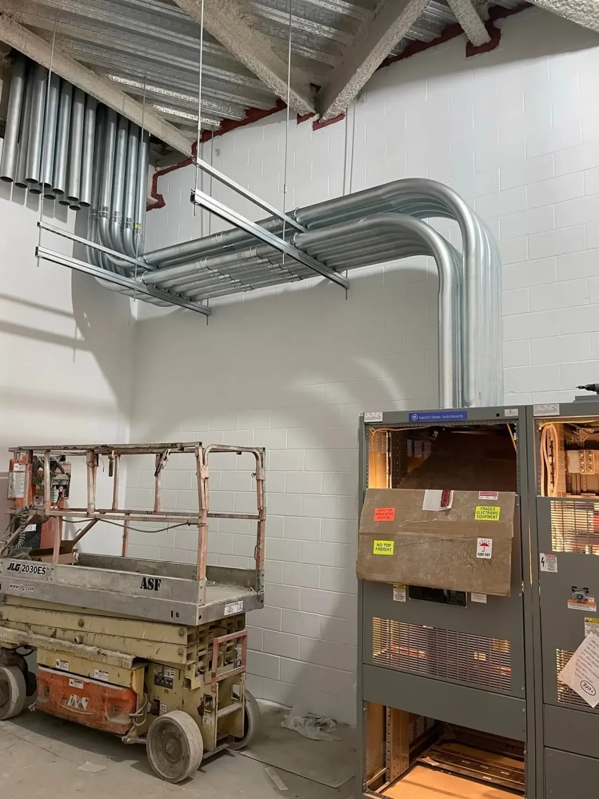 Indoor construction site with exposed ventilation pipes, a scissor lift, and electrical panels on a white brick wall.